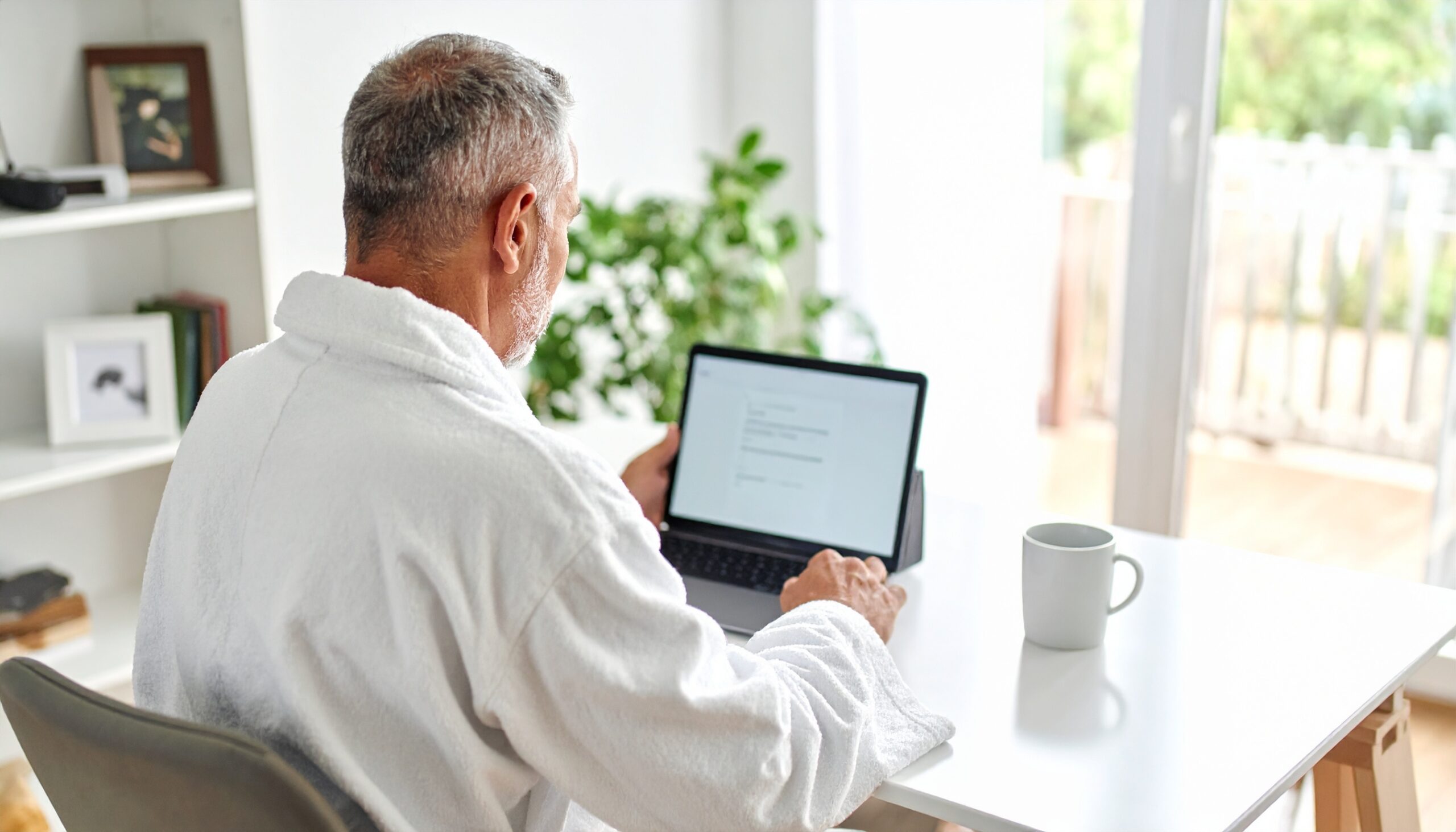 Senior male sitting at a home office desk, drinking a cup of coffee in his robe.